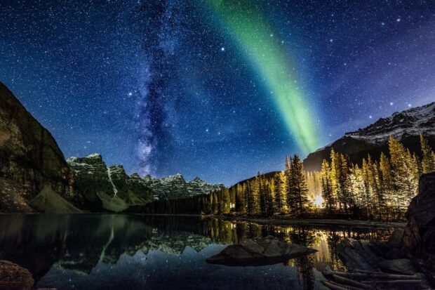 The starry sky above Moraine Lake with northern lights and pine trees illuminated at night