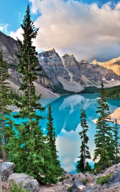 Tall evergreen trees near turquoise water at Moraine Lake surrounded by rocky mountains and clouds