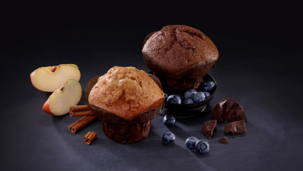 Two muffins with blueberries cinnamon sticks chocolate and apple slices on a dark surface