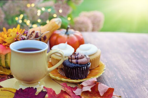 A chocolate muffin with two vanilla frosted cupcakes on a plate near a cup of tea and autumn leaves