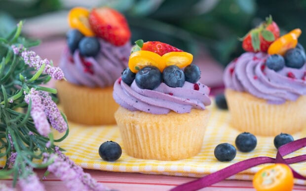 A cupcake with purple frosting decorated with fresh blueberries strawberry and kumquat on a yellow checkered cloth
