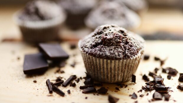 A close up of a chocolate muffin surrounded by chocolate pieces and crumbs on a wooden surface