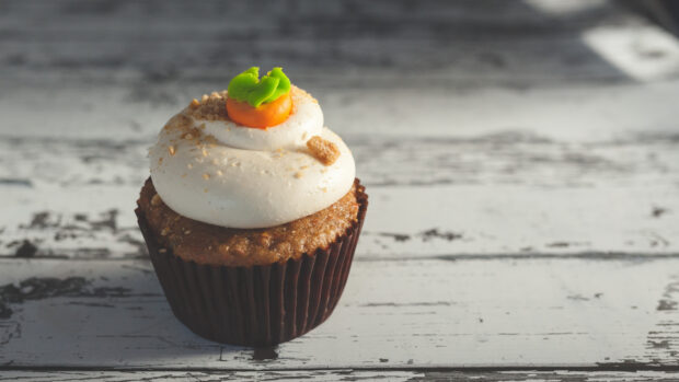A close up of a cupcake topped with creamy frosting and a small decorative carrot on a rustic wooden surface