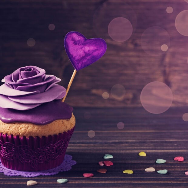 A muffin decorated with purple frosting and a flower shaped top with a purple heart on a stick on a wooden surface