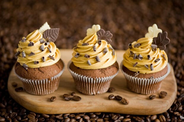 Three muffins with coffee flavored frosting and chocolate decorations on a wooden board
