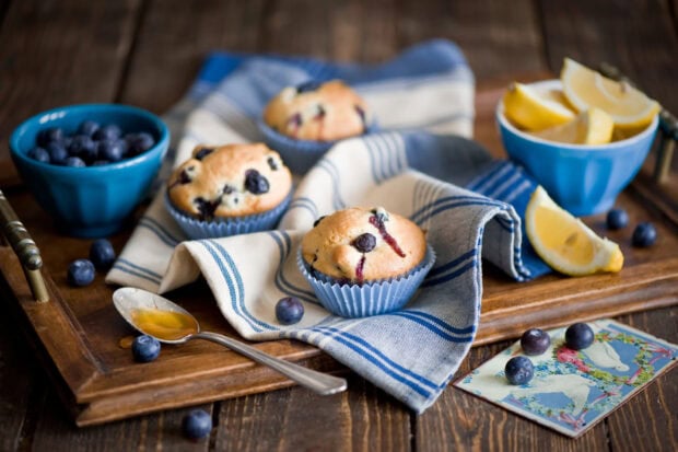 Blueberry muffins on a wooden tray with blueberries and lemon wedges on a cloth napkin