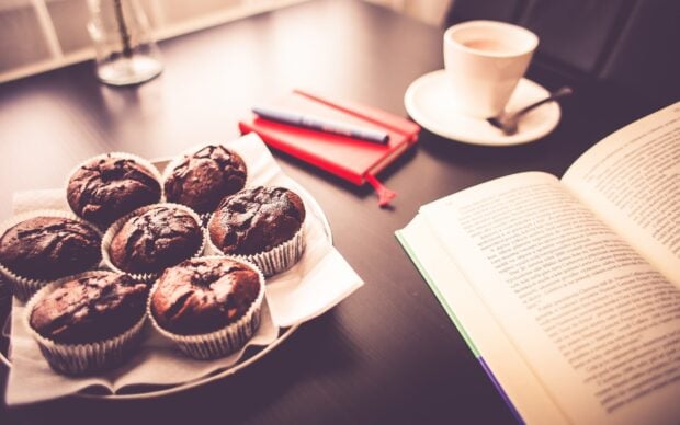 Chocolate muffins on a plate beside an open book and cup of coffee on a table