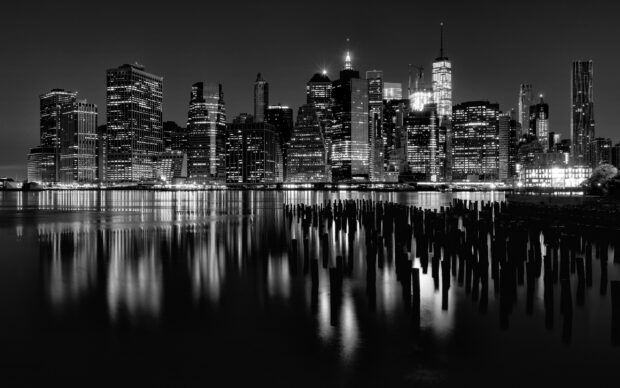 Black and white view of New York skyline reflecting on water with wooden pilings in the foreground