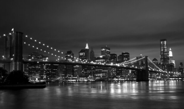 The New York skyline in black and white at night with the Brooklyn Bridge illuminated in the foreground
