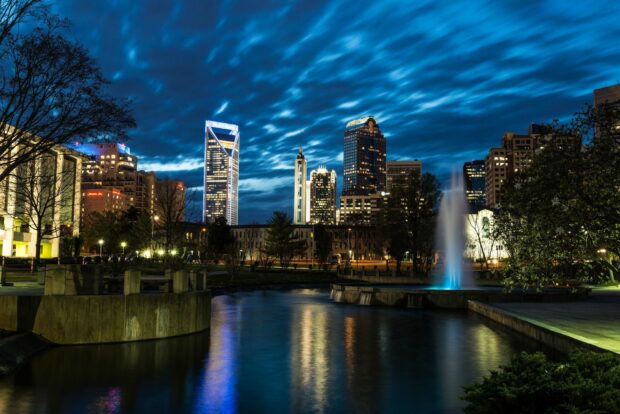 Nighttime cityscape of North Carolina with illuminated buildings and a water fountain reflecting on the lake