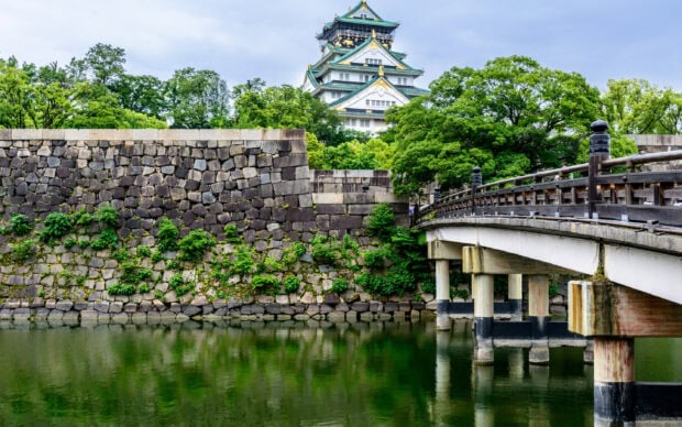A scenic view of Osaka Castle surrounded by lush greenery and stone walls along the river