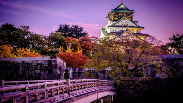 Osaka Castle surrounded by autumn trees and a wooden bridge at sunset with a purple sky