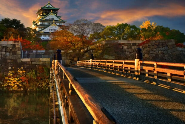 A scenic view of Osaka Castle with autumn trees and a wooden bridge at sunset