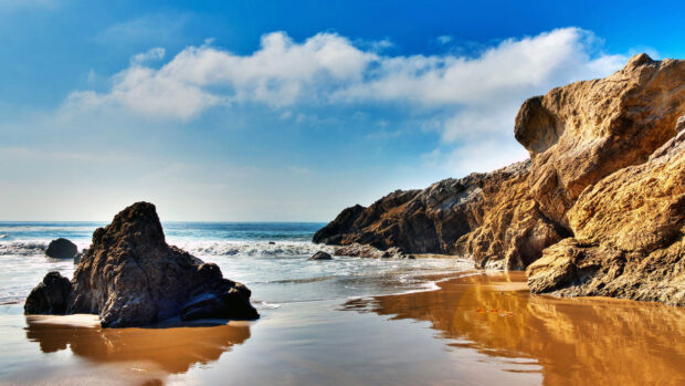 Rocky coastline under blue sky along the Pacific Ocean shore with wet sandy beach reflection