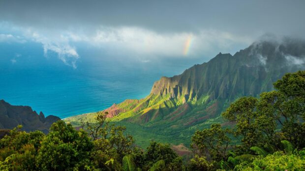 Lush green mountains overlooking the Pacific Ocean with a rainbow in the cloudy sky
