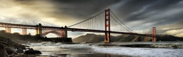 A scenic view of the Pacific coastline with a large suspension bridge and rocky shore under a dramatic sky