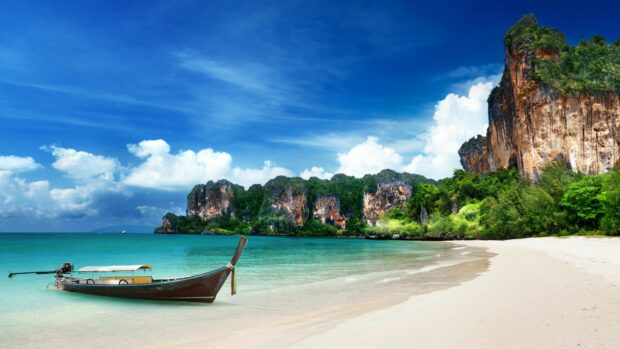 A traditional wooden longtail boat rests on clear turquoise water at Phi Phi with rocky cliffs and blue sky background