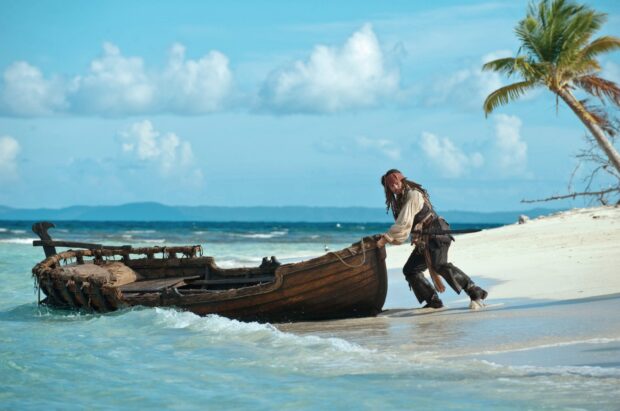 A pirate is pulling a wooden boat ashore on a tropical beach in Pirates Of The Caribbean