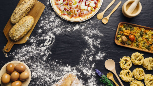 Fresh ingredients for pizza including eggs bread and pasta arranged on a black surface with flour scattered around