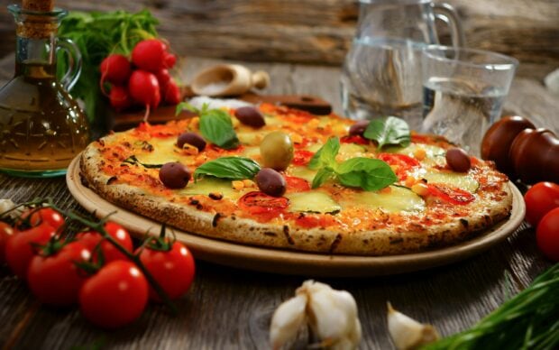 A delicious pizza with olives basil and tomatoes on a wooden table with garlic and olive oil