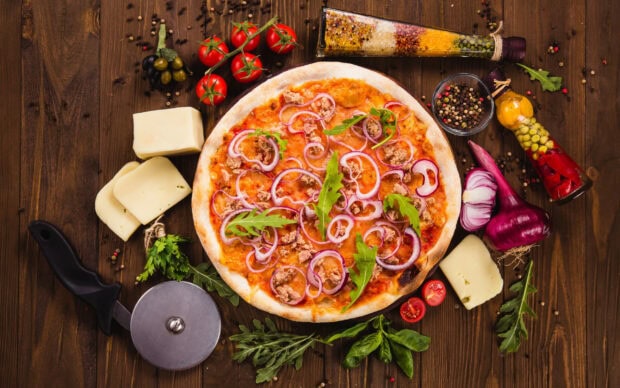 A delicious pizza topped with onions and greens surrounded by cheese and fresh ingredients on a wooden table