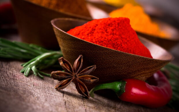 A close up of red powder and star anise with green herbs and chili pepper on wooden surface