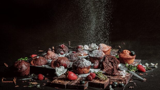 Chocolate muffins with berries being dusted with powdered sugar on a wooden board