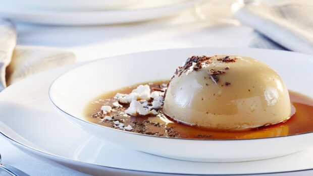 A creamy pudding dessert with chocolate shavings and sauce served on a white plate