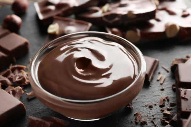 Smooth chocolate pudding in a glass bowl surrounded by chocolate pieces and nuts