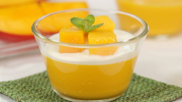 A glass cup of pudding topped with mango cubes and a mint leaf on a green mat