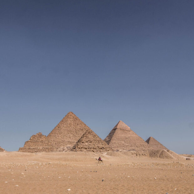 A camel rider crossing the desert near the ancient pyramids of Giza under a clear blue sky