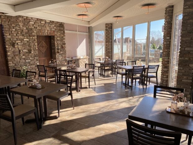 A sunlit restaurant interior with wooden tables and chairs near large glass windows featuring brick walls