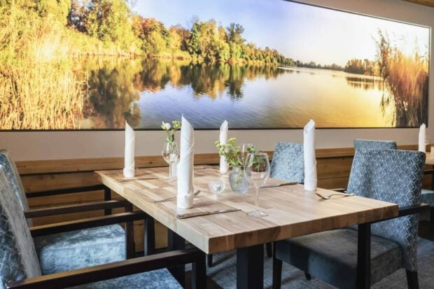 A restaurant table setting with elegant chairs and a nature scene wall art featuring trees and lake reflections