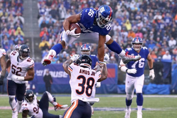 Saquon Barkley leaping over a defender during a football game with intense action and crowd in the background