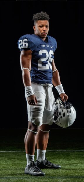 Saquon Barkley standing on grass field in football uniform holding a helmet