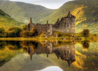 Historic Scottish castle ruins reflected in calm lake with green hills in the background