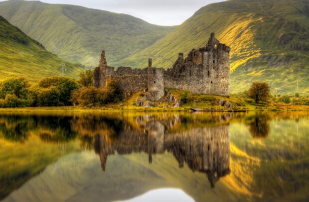 Historic Scottish castle ruins reflected in calm lake with green hills in the background