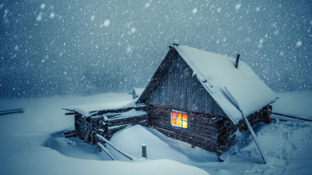 A wooden cabin covered in snow during snowfall with warm light glowing from the window