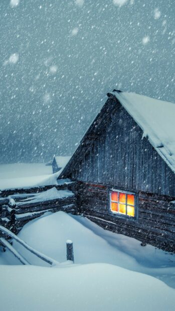 Snowfall surrounds a wooden cabin with warm light glowing through the window