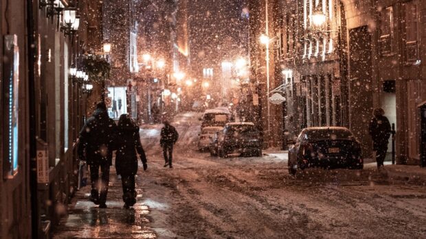 A snowy city street scene with snowfall and people walking along the sidewalks