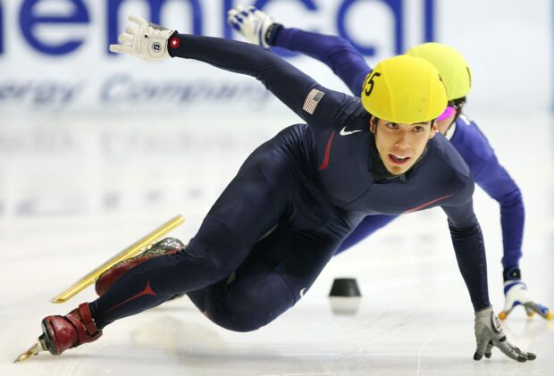 A speed skating athlete wearing a yellow helmet and navy suit racing on ice