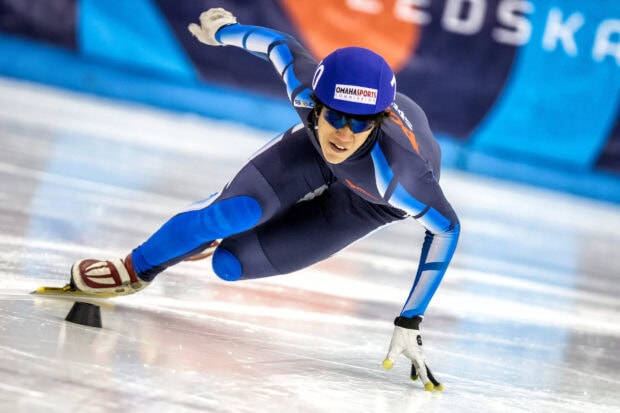 Male speed skating athlete leaning into a turn on the ice in a blue racing suit