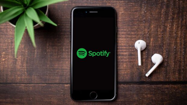 A phone screen displaying Spotify logo with wireless earbuds on wooden table
