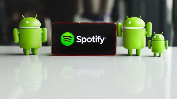 A smartphone displaying Spotify surrounded by multiple Android figurines reflecting on a white surface