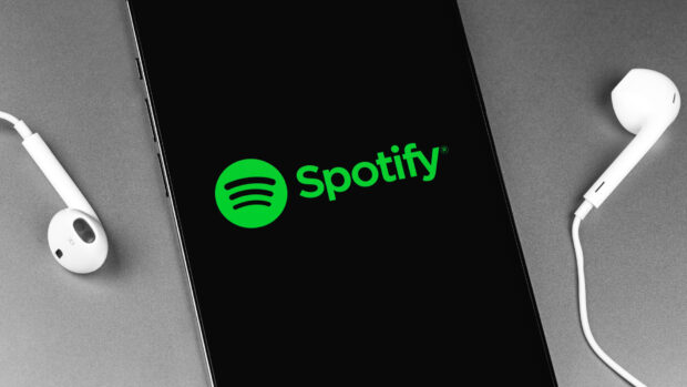 A smartphone displaying the Spotify logo with earphones placed beside it on a grey surface