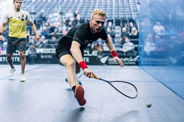 Male athlete lunging to hit a squash ball during a competitive match in a squash court