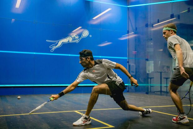 Two male athletes playing squash competing on a court with a blue wall and logos