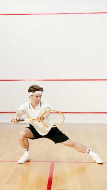 A male squash player in a white shirt and black shorts holding a racket while stretching on the court
