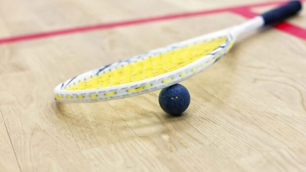 A squash racket with yellow strings resting on a blue squash ball on a wooden court floor