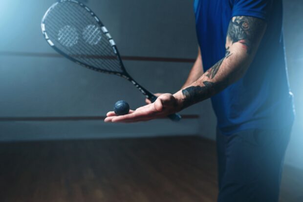 A tattooed player preparing to serve with a squash ball in hand on the court